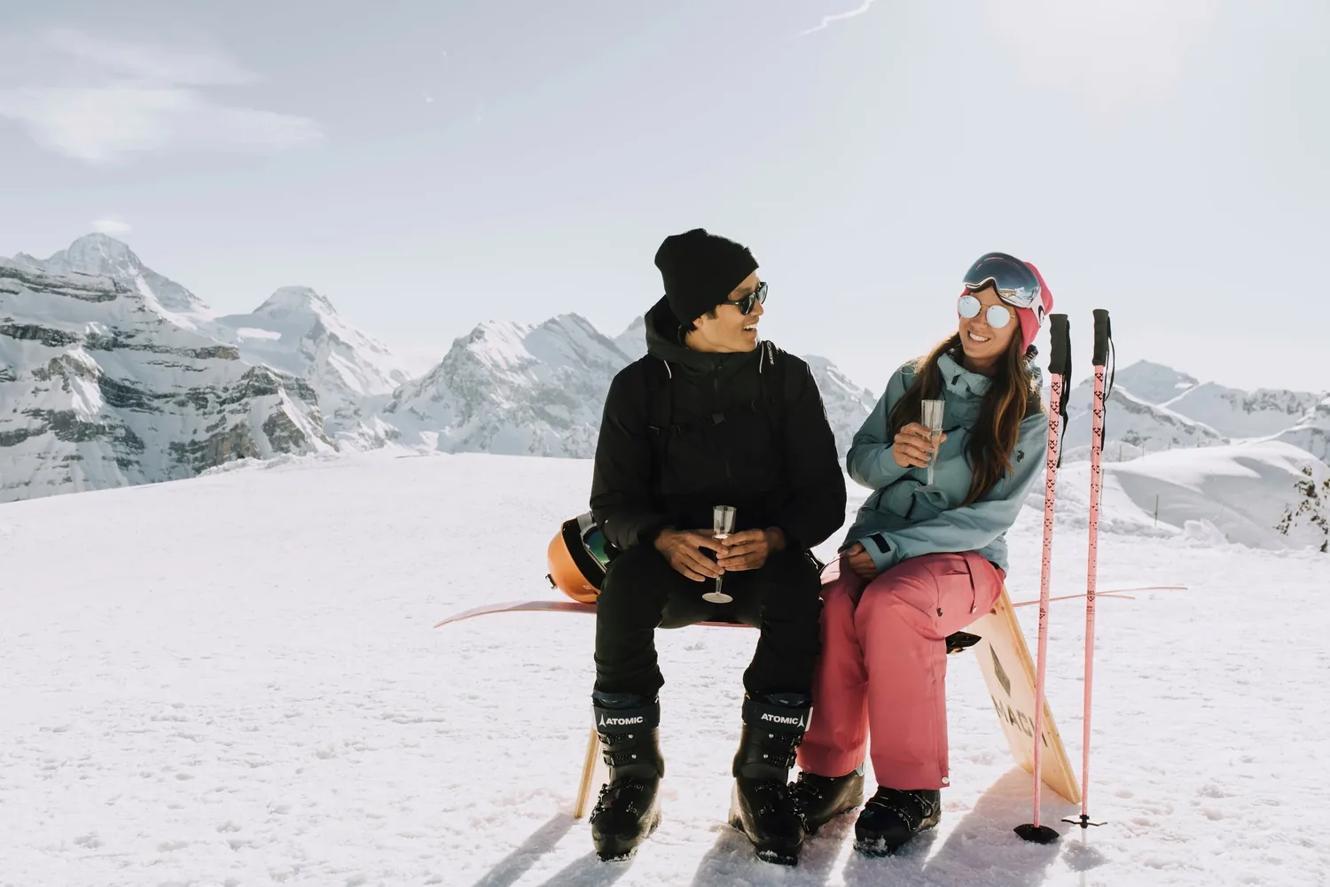 Alex and Vanessa celebrating with champagne on a ski slope, mountains behind them